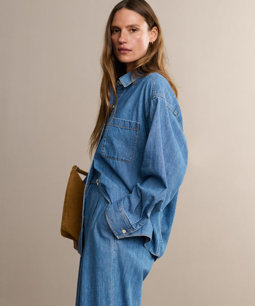 A woman with long brown hair stands sideways against a plain beige background, wearing the Parker Oversized Shirt with matching summer-weight denim pants and holding a tan clutch.