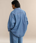 A person with long dark hair, wearing the Parker Oversized Shirt and matching denim pants, stands with their back to a beige background.