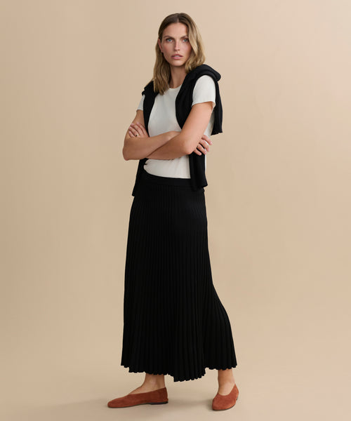 A woman with arms crossed wears a white t-shirt, black sweater over her shoulders, the long black Penelope Skirt, and brown flats against a plain beige background.