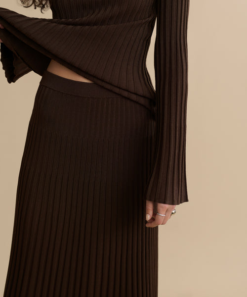 A person models the Penelope Skirt with a matching long-sleeve top, lifting the top to reveal the skirt's waistband. Only the torso and part of one arm are visible against a beige background.