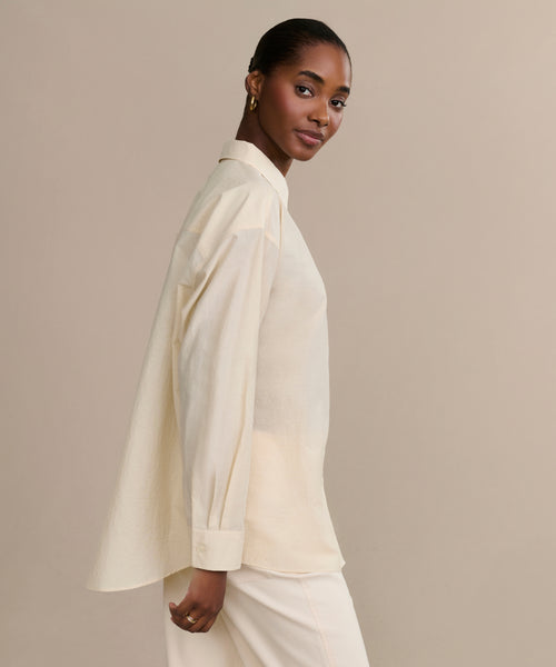 A woman stands sideways against a beige background, wearing the oversized Pierce Shirt with a collar and matching cream pants. Her hair is pulled back, and she faces the camera with a subtle smile.