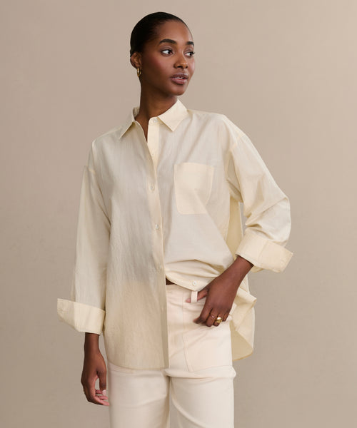 A woman with short, slicked-back hair and small hoop earrings poses confidently in a cream Pierce Shirt made of textured cotton, sleeves rolled up, paired with light pants, set against a plain beige background.