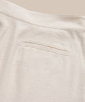 Close-up of Pleated Short in light beige textured fabric, high-waisted with a single welt back pocket and visible seam details, shown against a neutral background.