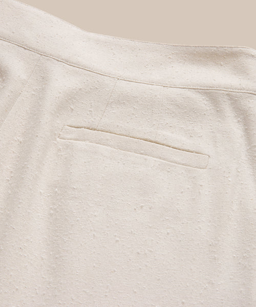 Close-up of Pleated Short in light beige textured fabric, high-waisted with a single welt back pocket and visible seam details, shown against a neutral background.