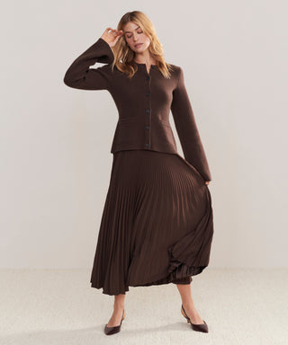 A woman models a Pleated Skirt with a brown buttoned cardigan and pointed heels, posing with one hand on her head against a plain light background.