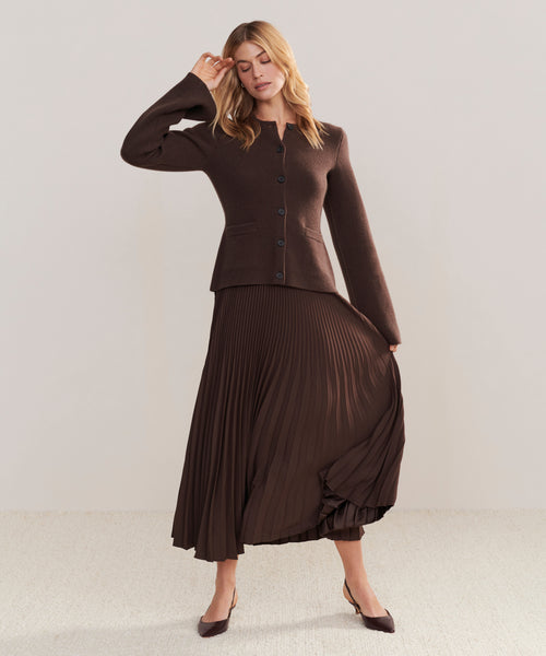 A woman models a Pleated Skirt with a brown buttoned cardigan and pointed heels, posing with one hand on her head against a plain light background.