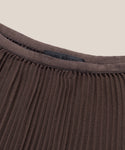 Close-up of the Pleated Skirt fabric in brown, featuring pleats and a smooth trim at the neckline with a faint black clothing label partially visible inside.