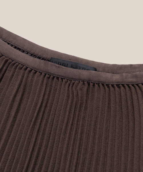 Close-up of the Pleated Skirt fabric in brown, featuring pleats and a smooth trim at the neckline with a faint black clothing label partially visible inside.