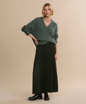 A woman with short blonde hair models a long black Pleated Skirt, pairing it with a loose green V-neck sweater, black boots, and a beaded necklace against a plain beige background.