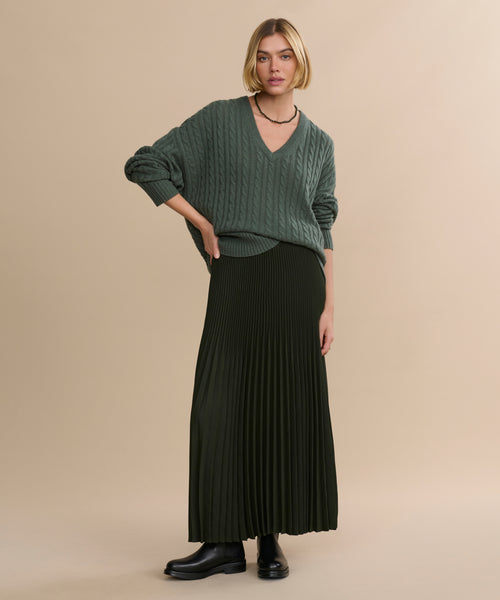 A woman with short blonde hair models a long black Pleated Skirt, pairing it with a loose green V-neck sweater, black boots, and a beaded necklace against a plain beige background.