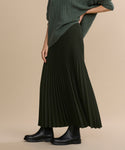 Wearing a dark green Pleated Skirt, a green cable knit sweater, and black ankle boots, this person stands against a beige background with their face not shown.