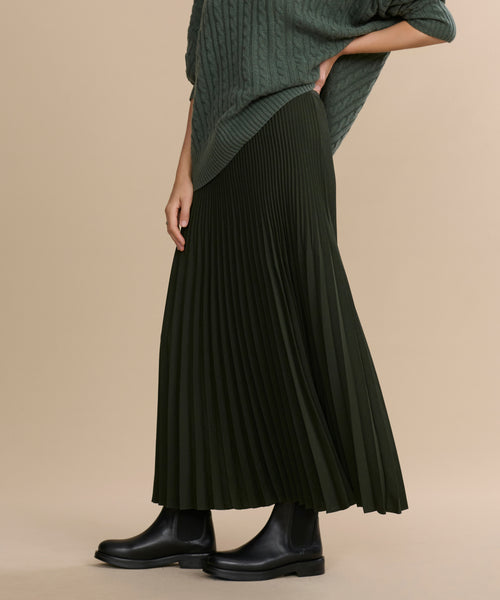 Wearing a dark green Pleated Skirt, a green cable knit sweater, and black ankle boots, this person stands against a beige background with their face not shown.