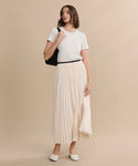 A woman with shoulder-length brown hair stands against a beige background, wearing an ivory Pleated Skirt, a white t-shirt, white flats, and holding a black shoulder bag while looking slightly to the side.
