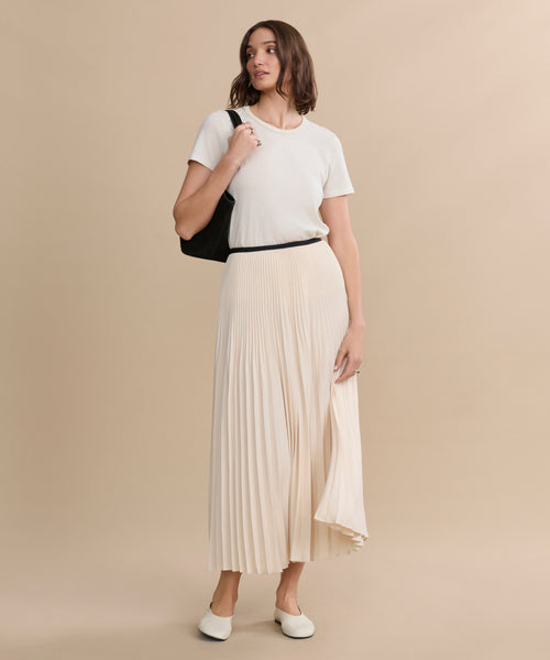 A woman with shoulder-length brown hair stands against a beige background, wearing an ivory Pleated Skirt, a white t-shirt, white flats, and holding a black shoulder bag while looking slightly to the side.