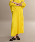 Someone wears the Pleated Skirt in bright yellow, paired with a matching sweater and black woven flats, standing arms crossed against a plain background.