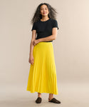 A woman with long curly hair smiles softly as she stands against a beige background, wearing a black top, black woven slip-on shoes, and the bright yellow matte Pleated Skirt with a flattering drape.