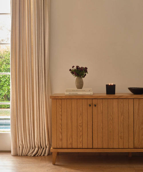 A wooden sideboard stands against a white wall near a window with light curtains. On the sideboard are stacked books, a Porta Ceramic Vase with purple flowers, a lit black candle, and a dark bowl—perfect for elegant home décor.