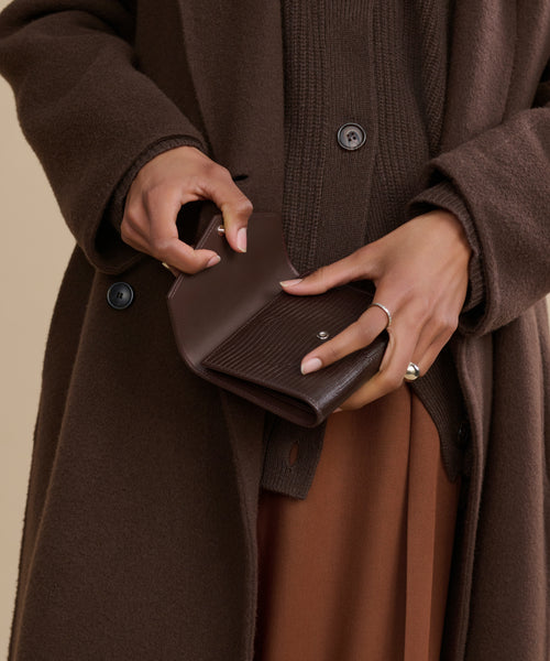 A person in a brown coat and sweater holds the Reece Leather Wallet, showing off neatly manicured hands and a silver ring on one finger.
