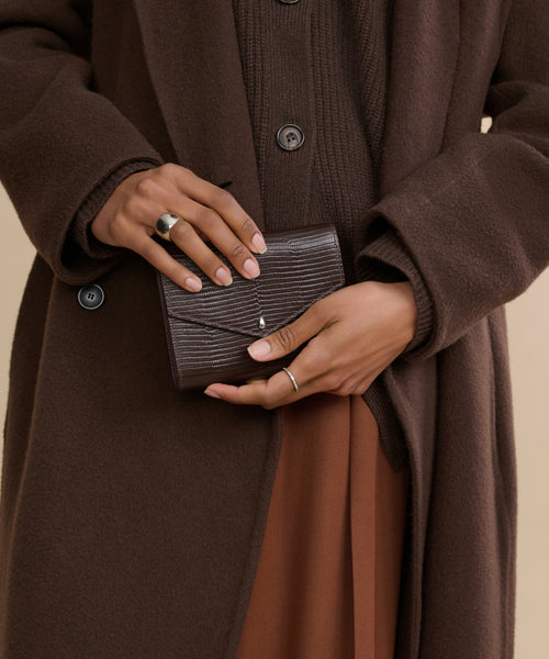A person wearing a brown coat and rust-colored skirt holds the Reece Leather Wallet with both hands, showing off its detailed embossing. Silver rings adorn their fingers, drawing focus to the wallet and their hands.
