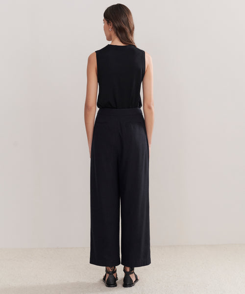 A woman faces away from the camera, wearing Relaxed Trouser in a wide-leg silhouette with a sleeveless black top and black sandals, set against a plain light background.