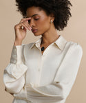 A woman with natural curly hair wears the Remy Shirt, a satin blouse with puffed sleeves, paired with silver hoop earrings. Standing against a beige background, she closes her eyes and gently touches her forehead in a thoughtful pose.