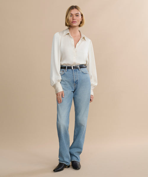 A woman with a blonde bob stands against a beige background, wearing the cream Remy Shirt with puffed sleeves, light blue high-waisted jeans, a black belt, and black shoes.