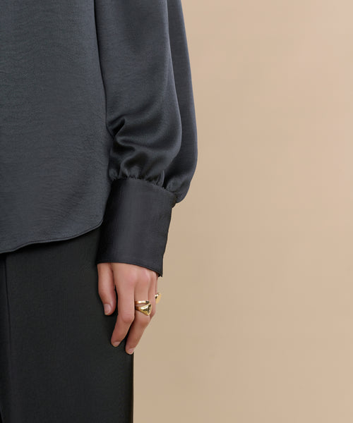 Wearing the Remy Shirt with puffed sleeves and black pants, a person stands against a beige background. Their hand, decorated with gold rings, rests by their side.