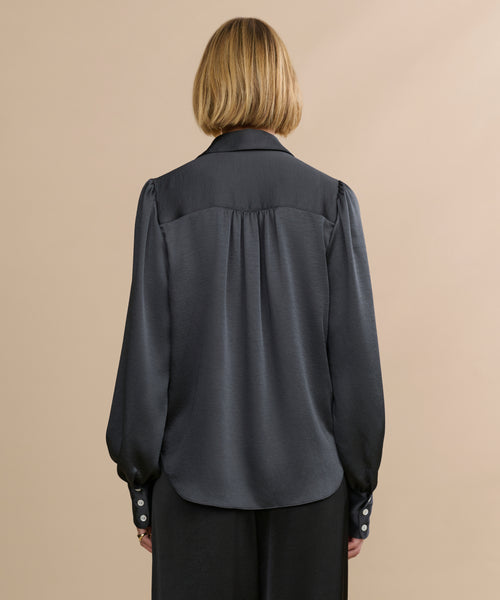 A person with blonde, chin-length hair is seen from behind wearing the Remy Shirt, featuring vintage-inspired satin fabric, puffed sleeves, and buttoned cuffs, paired with black pants against a plain beige background.