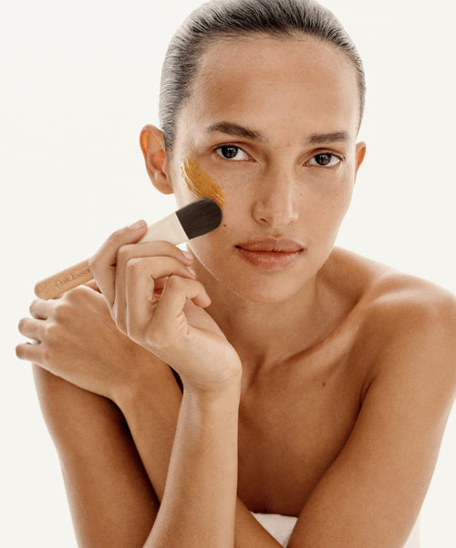A woman with bare shoulders applies Restorative Mask to her cheek with a brush. She looks directly at the camera, set against a white background.