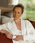 A woman relaxes indoors on a rust-colored sofa, wearing a white robe and the Restorative Mask on her face, holding its small jar and gazing thoughtfully to the side.