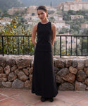 A woman wearing the sleeveless black Cotton Rib Tank and a long black skirt stands on a stone terrace with her hands behind her back, overlooking a scenic hillside village filled with stone buildings and lush greenery.