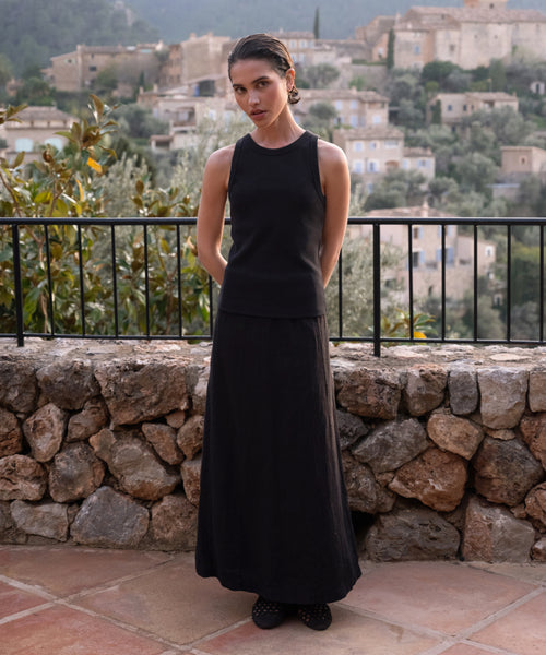 A woman wearing the sleeveless black Cotton Rib Tank and a long black skirt stands on a stone terrace with her hands behind her back, overlooking a scenic hillside village filled with stone buildings and lush greenery.