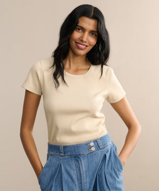 A woman with long, wavy black hair smiles while wearing the Rib Tee and high-waisted blue pleated jeans, standing against a plain light tan background.