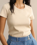 A woman wears a short-sleeved beige Rib Tee with a U-shaped neck, tucked into high-waisted blue jeans with pleats and visible buttons. Her face is partially out of frame.