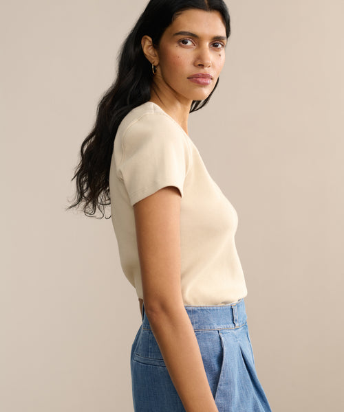 A woman with long dark hair wears the Rib Tee and high-waisted blue pants, standing sideways and looking at the camera against a plain beige background.