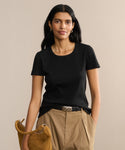 A woman with long dark hair wears the Rib Tee—a black fitted top with a U-shaped neckline—paired with tan high-waisted pants and a matching handbag, standing against a beige background and smiling slightly at the camera.