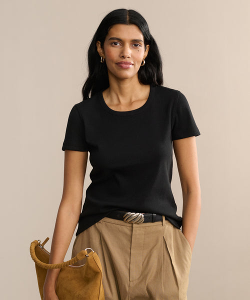 A woman with long dark hair wears the Rib Tee—a black fitted top with a U-shaped neckline—paired with tan high-waisted pants and a matching handbag, standing against a beige background and smiling slightly at the camera.
