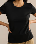 A woman models the black Rib Tee, featuring a fitted silhouette and U-shaped neckline, paired with beige pants. She stands hands on hips against a plain background, face not visible.