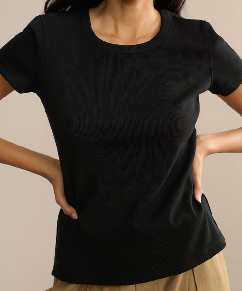 A woman models the black Rib Tee, featuring a fitted silhouette and U-shaped neckline, paired with beige pants. She stands hands on hips against a plain background, face not visible.