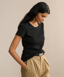 A woman with long dark hair wears the Rib Tee—a black, fitted top with a U-shape neck—paired with tan pleated pants. She stands against a beige background, hands in pockets, looking down with a slight smile.