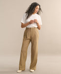 A woman with long curly hair wears a white tee and lightweight linen Riley Trouser, paired with cream shoes. She stands against a plain beige background, looking down with her hands together.