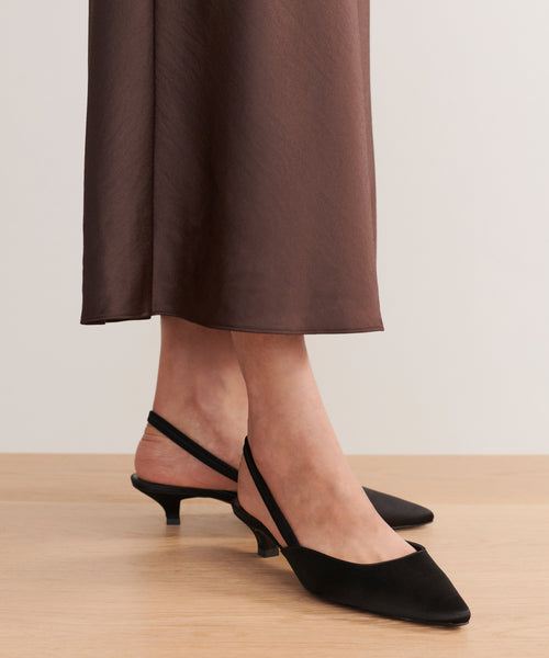 A person wearing a brown skirt and elegant Satin Kitten Heel shoes with a low heel and pointed toe stands on a wooden floor. This made-in-Italy footwear highlights refined style.