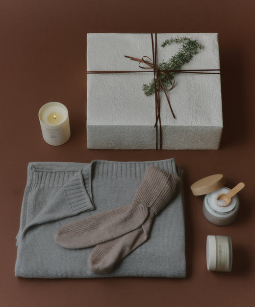 A cozy flat lay displays a folded gray sweater, beige socks, a lit candle, a cream jar with spatula, and the Seasonal Essentials Gift Box tied with string and greenery, all arranged on a warm brown background.