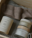 The Seasonal Essentials Gift Box includes a beige sock, grey knitwear, a Jenni Kayne Cedar candle, and two Oak Essentials skincare items in a wooden box—the perfect self-care holiday gift set.