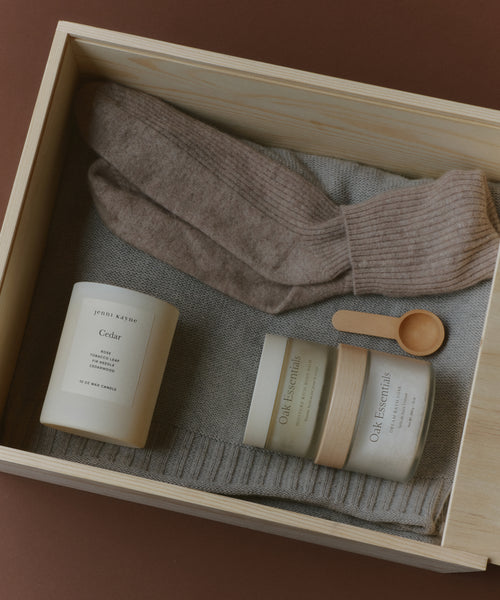 The Seasonal Essentials Gift Box features a wooden box with beige wool socks, a gray knit sweater, a white cedar-scented candle, two Oak Essentials skincare jars, and a small wooden spoon—all elegantly arranged.