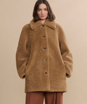 A woman with wavy brown hair stands against a plain beige background, wearing the Shearling Aurora Coat—a light brown, plush shearling coat with large buttons—paired with brown pants.