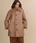 A woman models the Shearling Aurora Coat, featuring a wide collar and large buttons, with her hands in her pockets. Set against a beige backdrop, she highlights the cozy and timeless charm of this reversible tan coat.