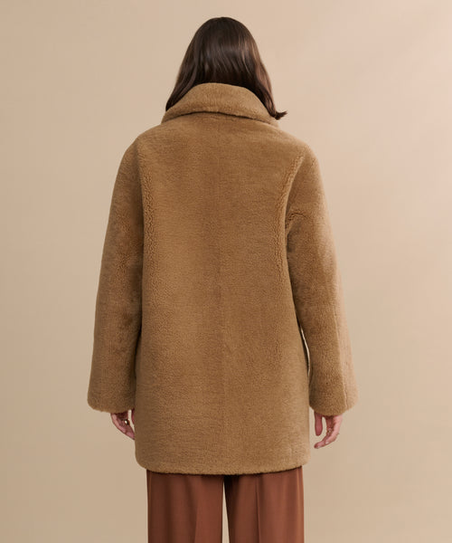 A person with long brown hair is seen from behind, wearing the Shearling Aurora Coat in light brown and brown pants, standing against a neutral beige background.