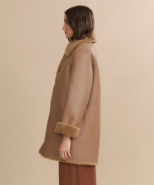 A woman in profile wears the Shearling Aurora Coat, featuring a fur-lined collar and cuffs, paired with brown pants. Her chin-length wavy hair frames her face against a plain beige background.