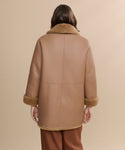A person with medium-length brown hair, seen from behind, wears the Shearling Aurora Coat with turned-up cuffs and matching brown pants against a beige background.